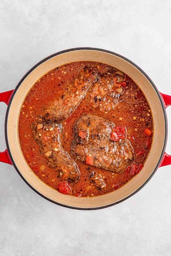 Red pot with cooked beef steaks in a rich tomato-based sauce.