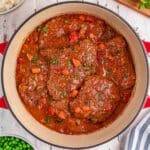 A pot of swiss steaks with carrots, tomatoes, and herbs in a red sauce.