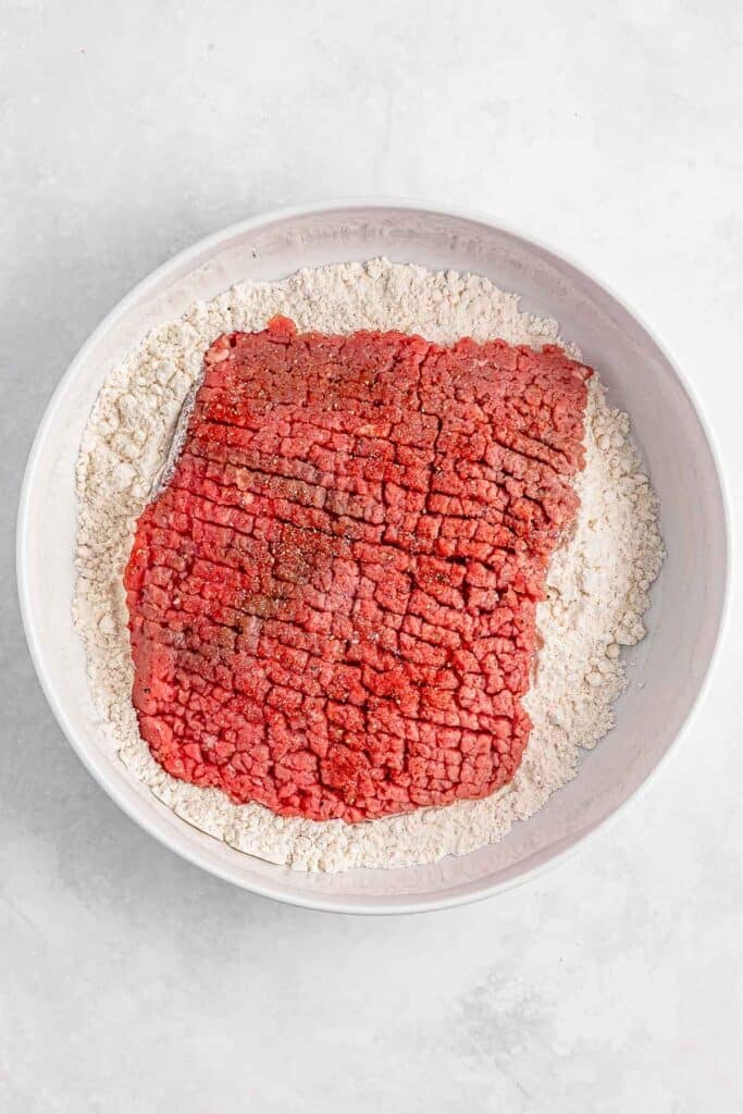 A piece of raw meat coated with flour in a white bowl.