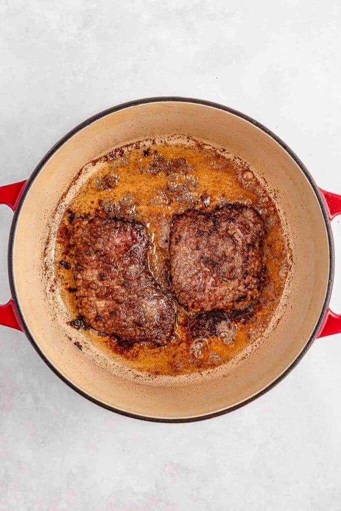 Two browned beef patties frying in a round red pan with sizzling oil.