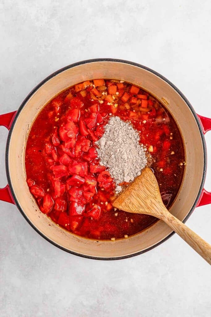A red pot with diced tomatoes, beef broth, and brown gravy mix being added to carrot and onions.