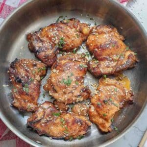 Cooked miso chicken thighs topped with sesame seeds and herbs in a skillet on a red and white checkered cloth.