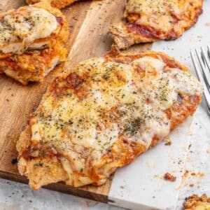 Close-up of air fryer chicken parmesan topped with melted cheese and herbs on a wooden board, with a fork nearby.