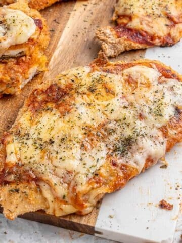 Close-up of air fryer chicken parmesan topped with melted cheese and herbs on a wooden board, with a fork nearby.