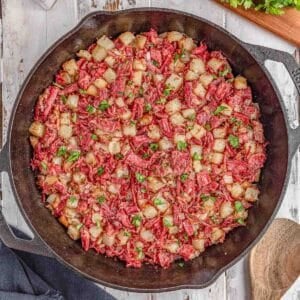 A cast iron skillet filled with corned beef hash, featuring diced potatoes, chopped corned beef, and garnished with parsley on a rustic wooden surface.
