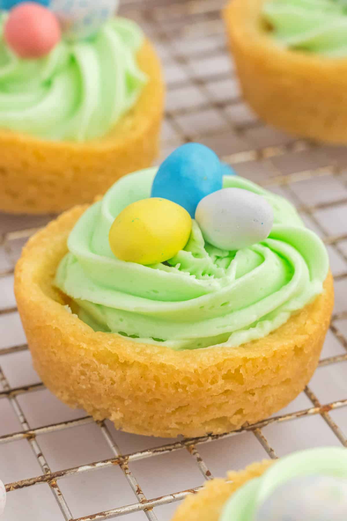 Easter basket cookie cup with green frosting and three small pastel colored candy eggs on top, resting on a wire rack.