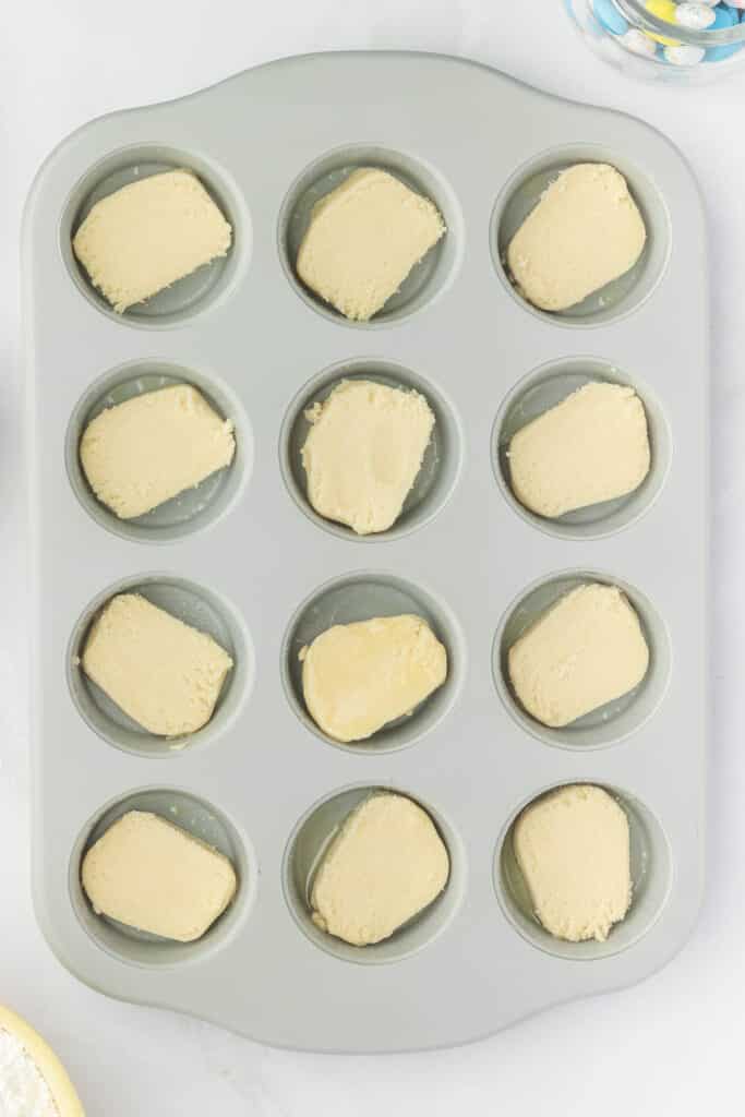 Muffin tray with sections filled with uncooked cookie dough placed on a light surface.