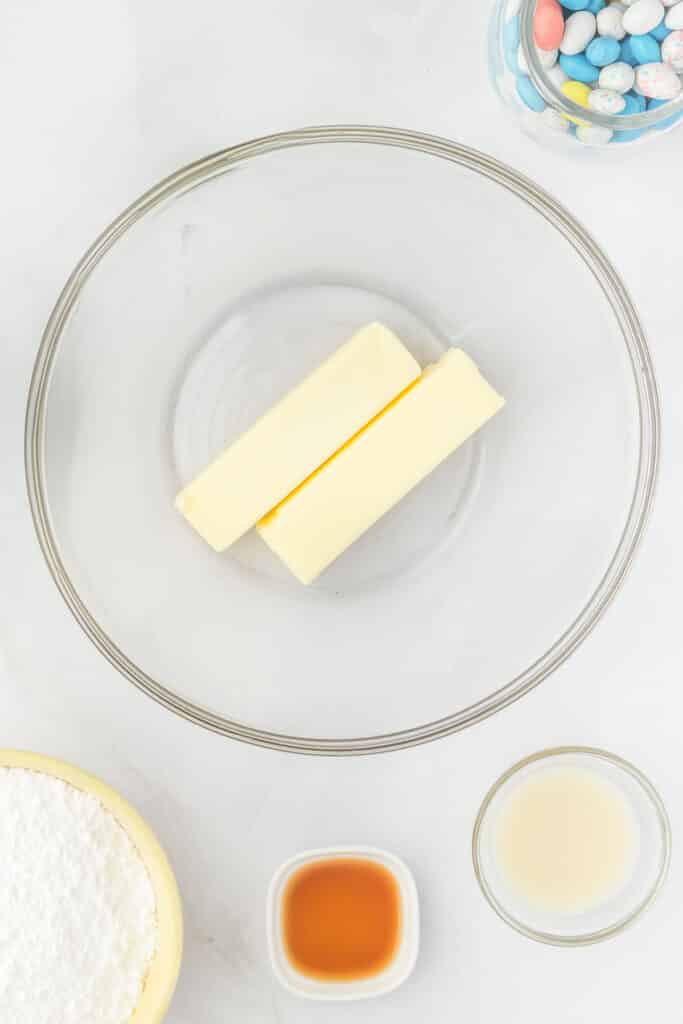Two sticks of butter in a clear mixing bowl, surrounded by small bowls containing vanilla extract, lemon juice, powdered sugar, and a jar of pastel-colored candy eggs.