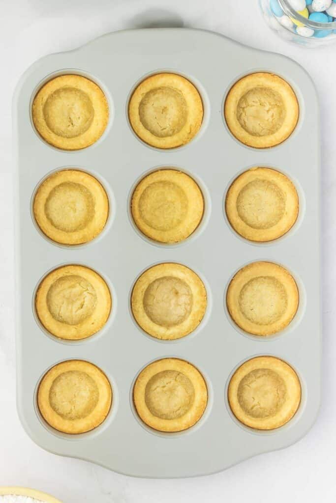 A muffin tray with twelve baked cookie cups, each with a golden-brown color, on a light gray surface.