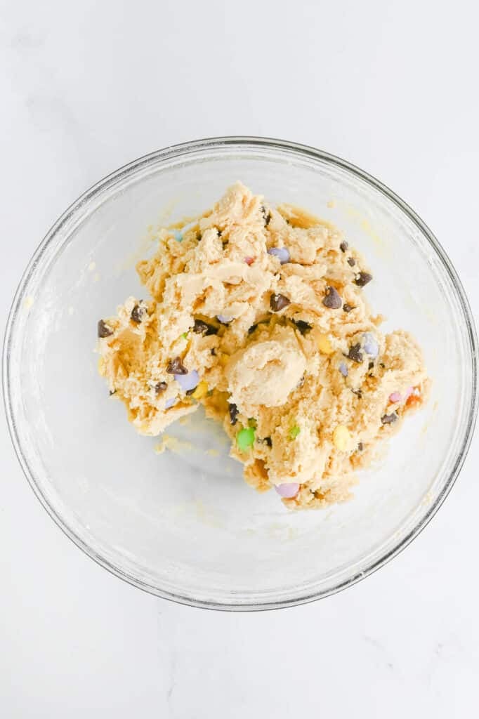 A glass mixing bowl with cookie dough mixed with chocolate chips and colorful M&M candy pieces on a white surface.
