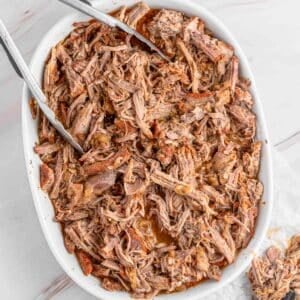 A close up of a white dish filled with instant pot pulled pork, with metal tongs placed on top.
