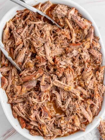 A close up of a white dish filled with instant pot pulled pork, with metal tongs placed on top.
