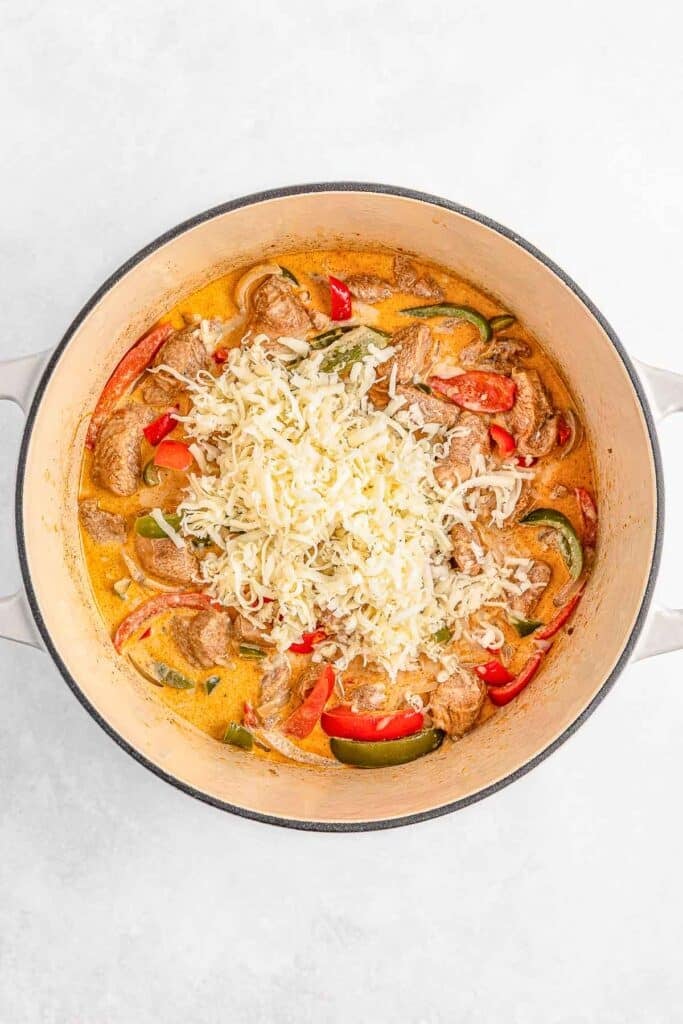 A pot of creamy soup with sliced peppers and onions, chicken, and a pile of grated cheese on top.