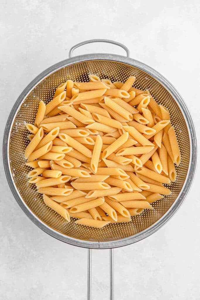 Cooked penne pasta draining in a metal colander on a light background.