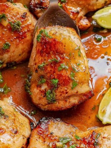 Close-up of cooked cilantro lime chicken thighs recipe in a flavorful sauce with herbs and lime wedges, served in a pan.