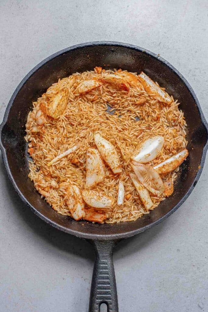 A cast iron skillet containing uncooked long rice, sliced onions and garlic with spices.