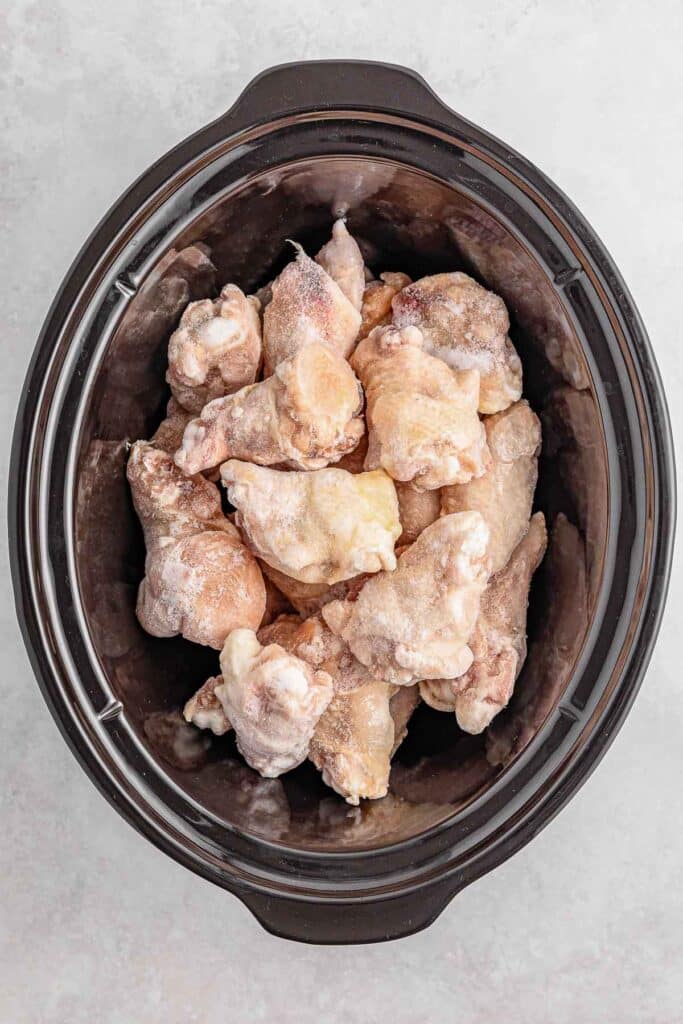 Frozen chicken wings placed inside a slow cooker on a light background.