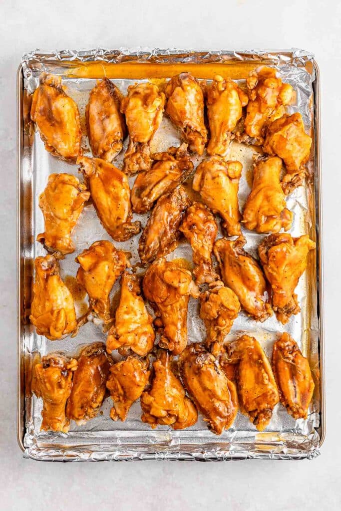 A baking tray lined with foil holds several baked chicken wings coated in a hot honey sauce.