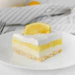 A slice of lemon lush dessert with creamy layers and a thin crust, topped with whipped cream and two lemon slices, on a white plate.