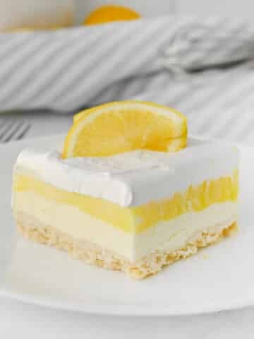 A slice of lemon lush dessert with creamy layers and a thin crust, topped with whipped cream and two lemon slices, on a white plate.