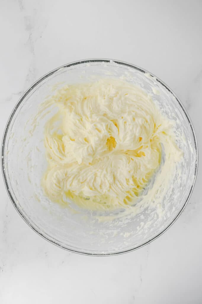 A glass bowl filled with cream cheese frosting on a white marble surface.