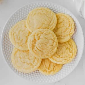 A white plate with seven round lemon sugar cookies on a white surface.