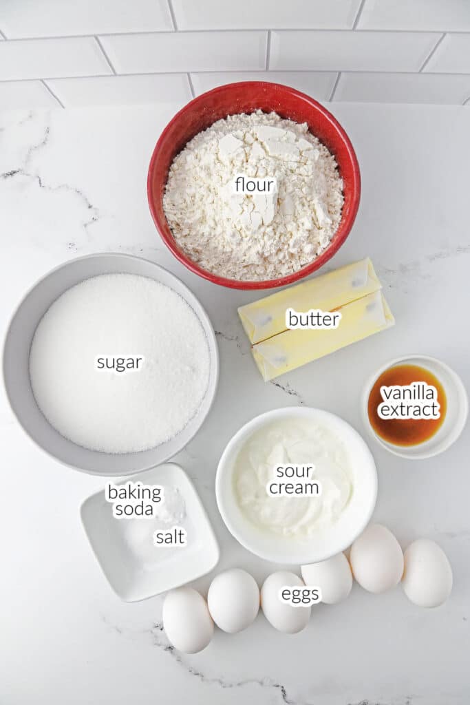 Top-down view of labeled baking ingredients for Sock it to me cake including: flour, butter, sugar, baking soda, salt, sour cream, vanilla extract, and eggs, arranged on a white surface.