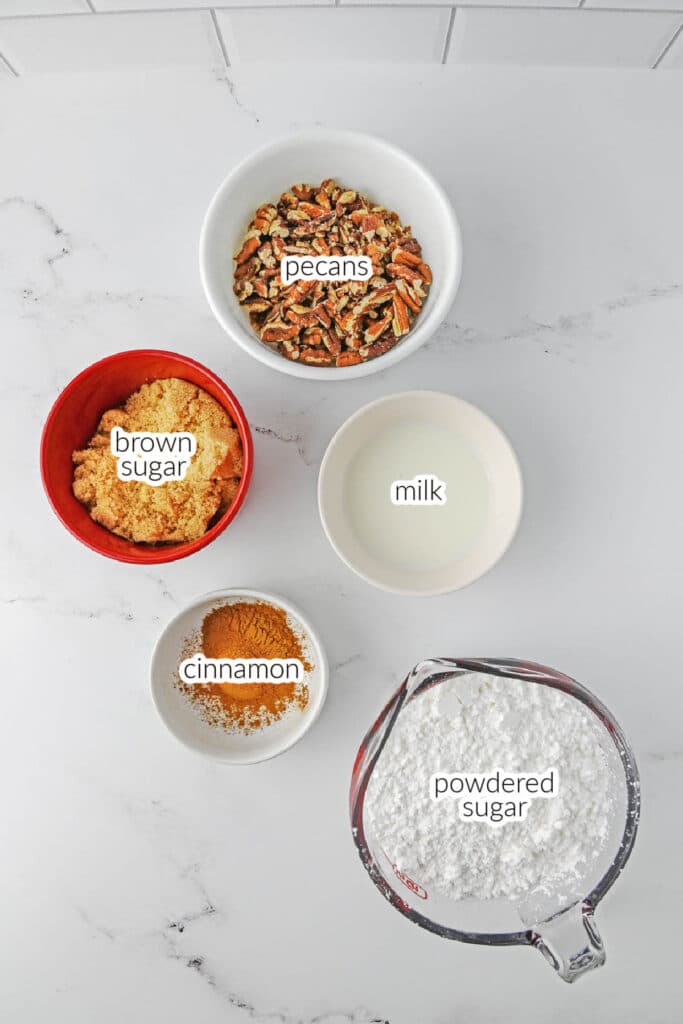 Ingredients for Sock it to me cake in five labeled bowls and a measuring cup containing pecans, brown sugar, cinnamon, milk, and powdered sugar arranged on a white marble countertop.