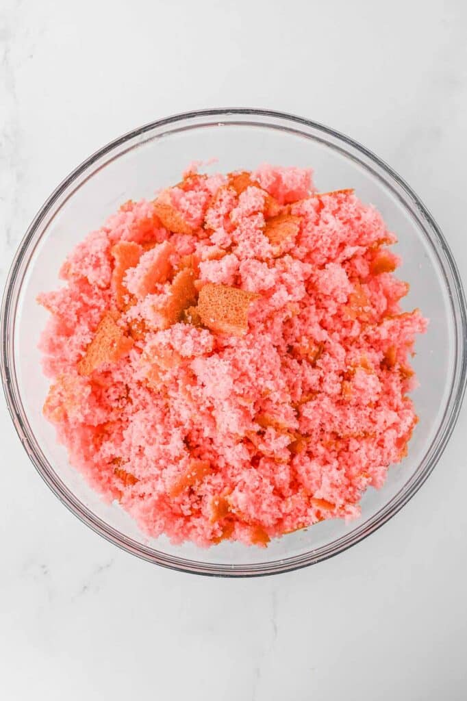 A clear glass bowl containing crumbled strawberry cake placed on a white marble surface.