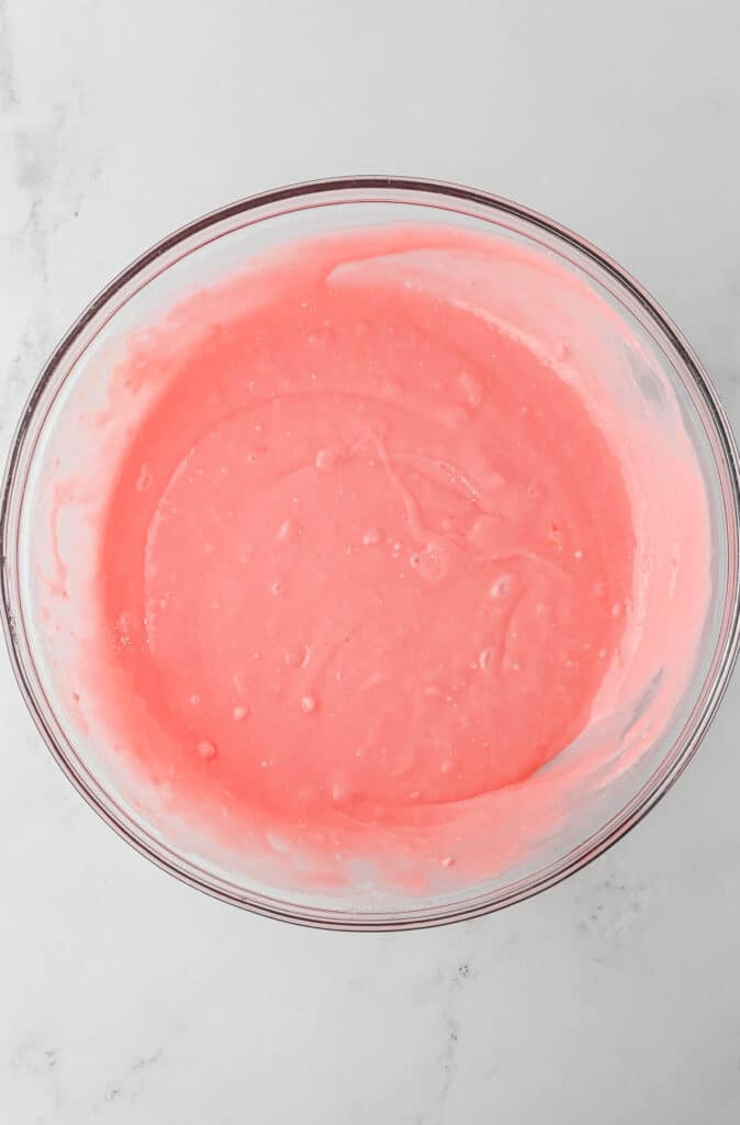 A glass bowl filled with strawberry cake batter rests on a white marble surface.