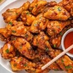A plate of glazed hot honey chicken wings garnished with herbs, served with a small bowl of honey with a wooden spoon.
