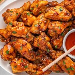 A plate of glazed hot honey chicken wings garnished with herbs, served with a small bowl of honey with a wooden spoon.