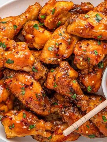 A plate of glazed hot honey chicken wings garnished with herbs, served with a small bowl of honey with a wooden spoon.