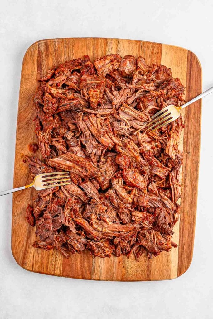 Shredded cooked beef on a wooden cutting board, two forks placed on opposite sides for pulling apart the meat.