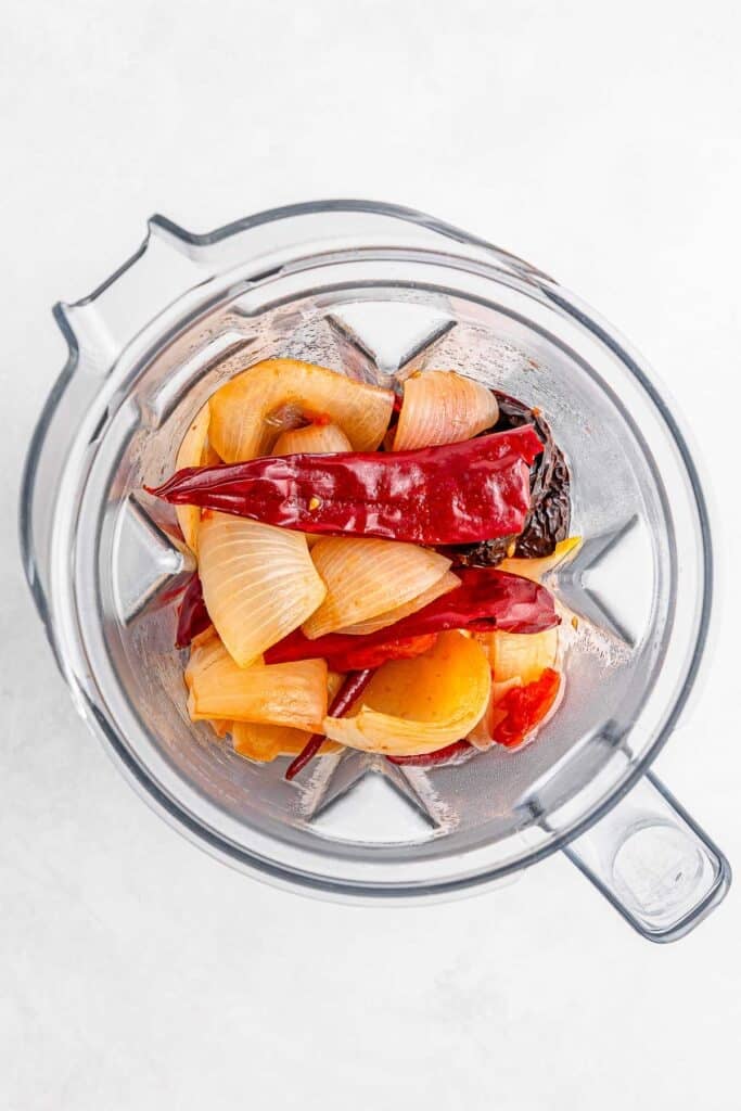 A blender with tomatoes, onions, garlic cloves, and chiles in it.