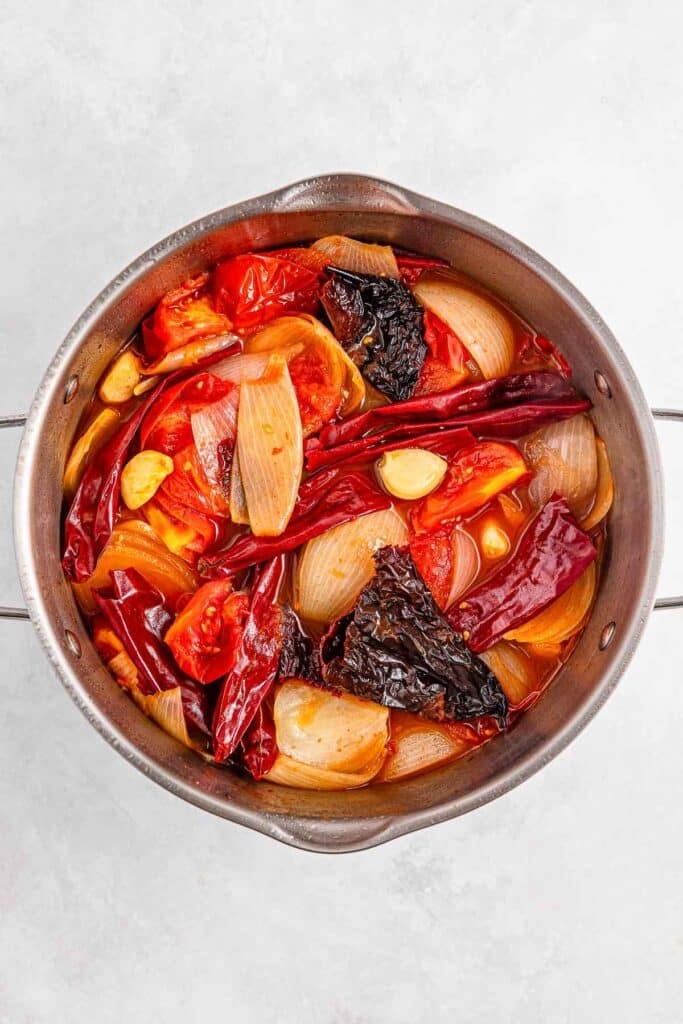 A stainless steel pot filled with chopped onions, tomatoes, chilies, and garlic cloves in broth, viewed from above.