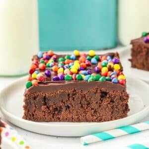 A square slice of cosmic brownie topped with chocolate ganache and rainbow chocolate chips on a white plate.