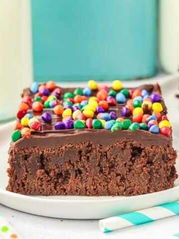 A square slice of cosmic brownie topped with chocolate ganache and rainbow chocolate chips on a white plate.