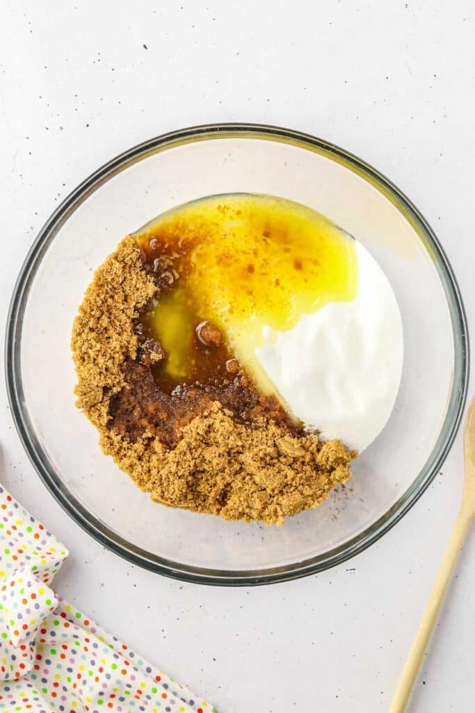 A glass bowl containing brown sugar, granulated sugar and melted butter with a wooden spoon.