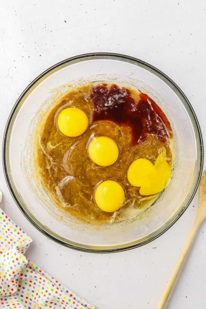 A glass mixing bowl contains four cracked eggs and vanilla extract mixed into the sugar mixture.