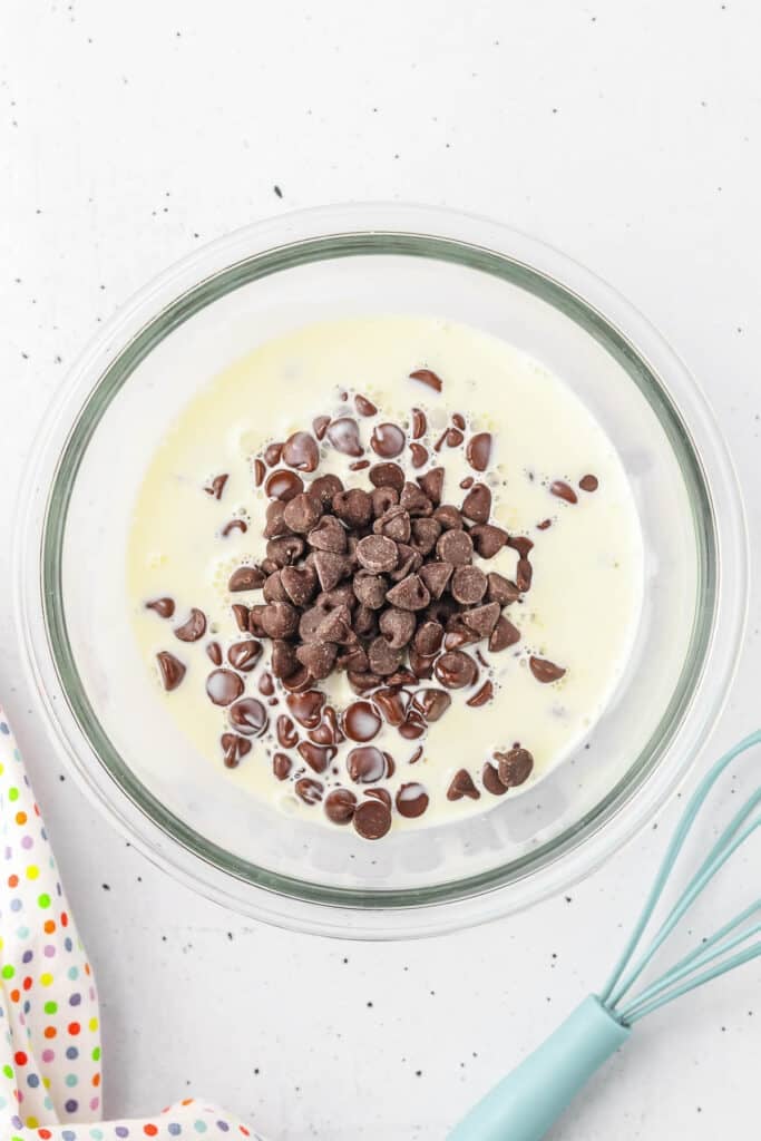 A glass bowl containing heavy cream and a pile of chocolate chips.