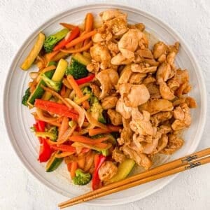 A white plate with hibachi chicken and a mix of sautéed vegetables including carrots, broccoli, zucchini, and red and orange bell pepper with chopsticks on the side.