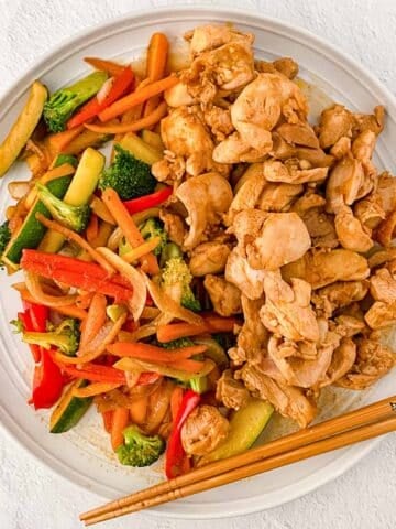 A white plate with hibachi chicken and a mix of sautéed vegetables including carrots, broccoli, zucchini, and red and orange bell pepper with chopsticks on the side.