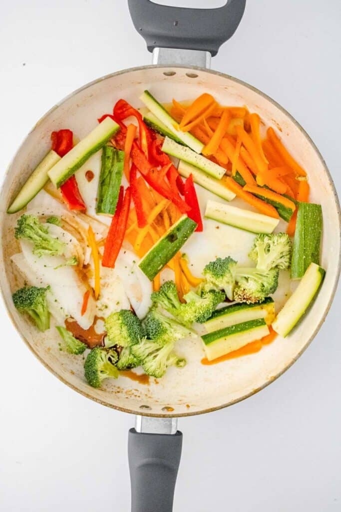 A white pan containing sliced zucchini, red and orange bell pepper, carrot, onion, and broccoli on a white background.