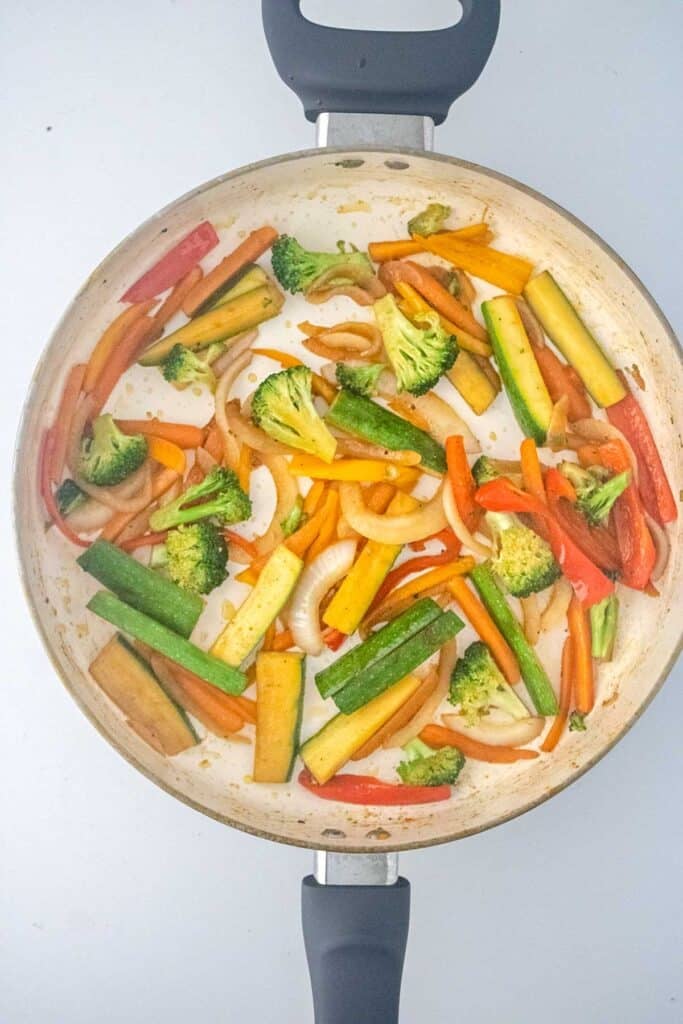 A white pan containing sauteed zucchini, red and orange bell pepper, carrot, onion, and broccoli on a white background.