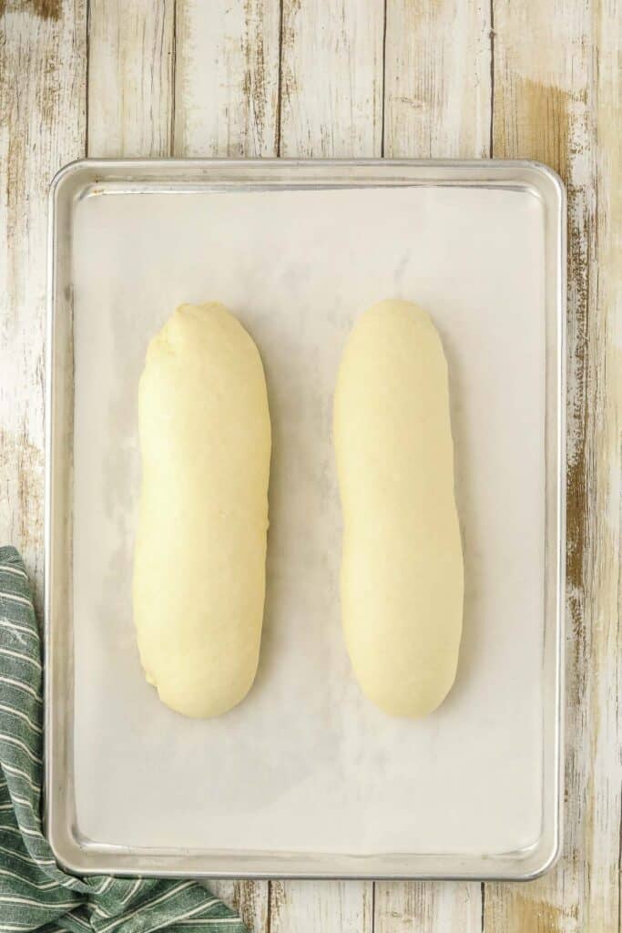 Two rolled, unbaked loaves of bread dough on a parchment-lined baking sheet.