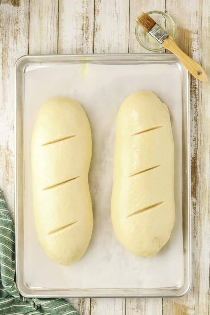 Two unbaked loaves of dough with three diagonal scores each rest on a parchment-lined baking sheet.