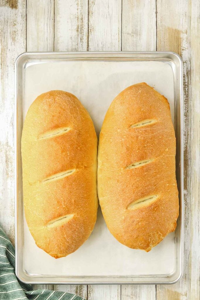Two loaves of golden-brown bread resting on a parchment-lined baking sheet on a wooden surface.