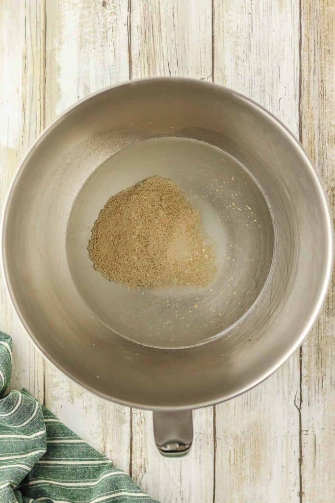 A metal mixing bowl containing dry yeast, water and sugar ready to be mixed together.