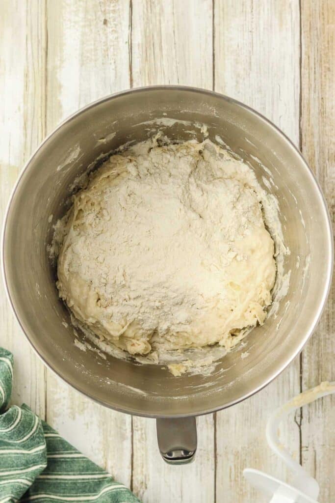 A metal mixing bowl contains bread dough with a layer of flour on top.